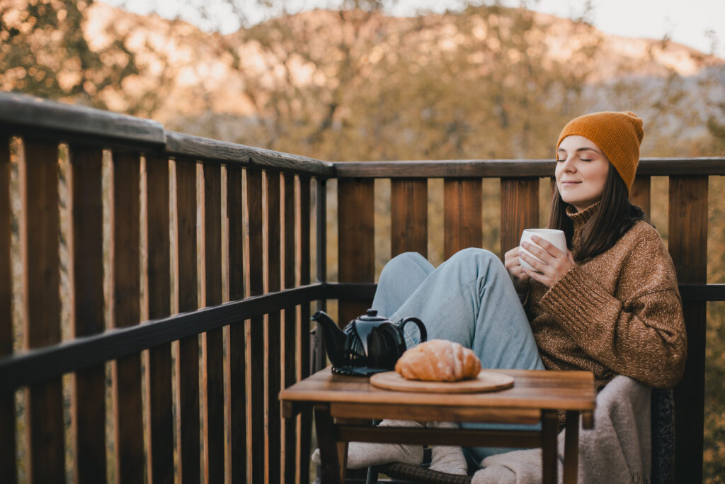 Weiblich gelesene Person sitzt im Herbst auf dem Balkon mit einer Tasse Tee in der Hand und einem Croissant auf einem Beistelltisch. Sie hat die Augen geschlossen und genießt den Moment.